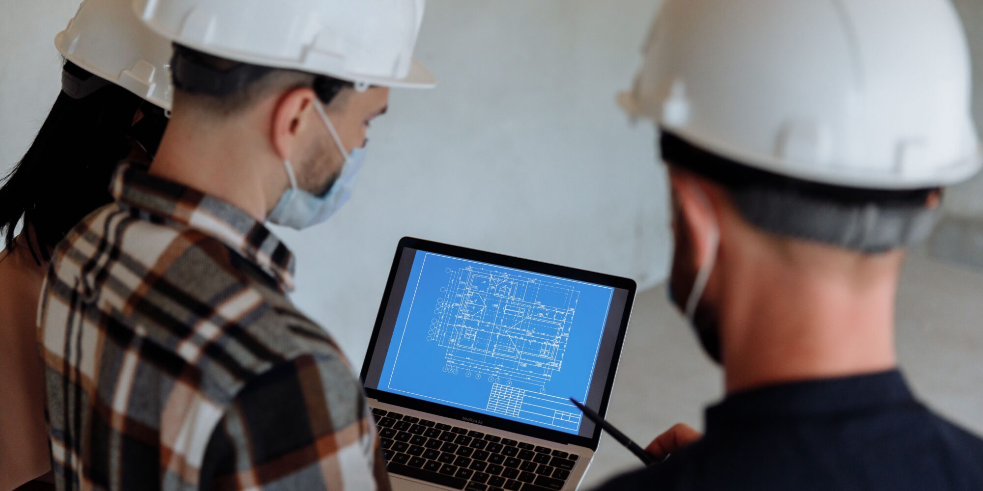 Tre byggprojektledare med vita hjälmar granskar en digital CAD-ritning på en bärbar dator ute på en byggarbetsplats.