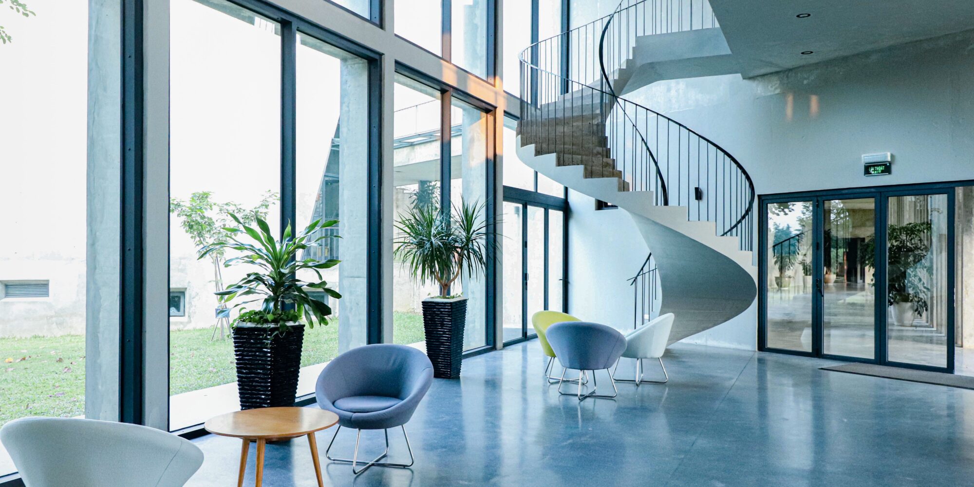 Ett högblankt, förädlat betonggolv i en modern kontorslounge med stora glaspartier, designmöbler och en spiraltrappa i betong.