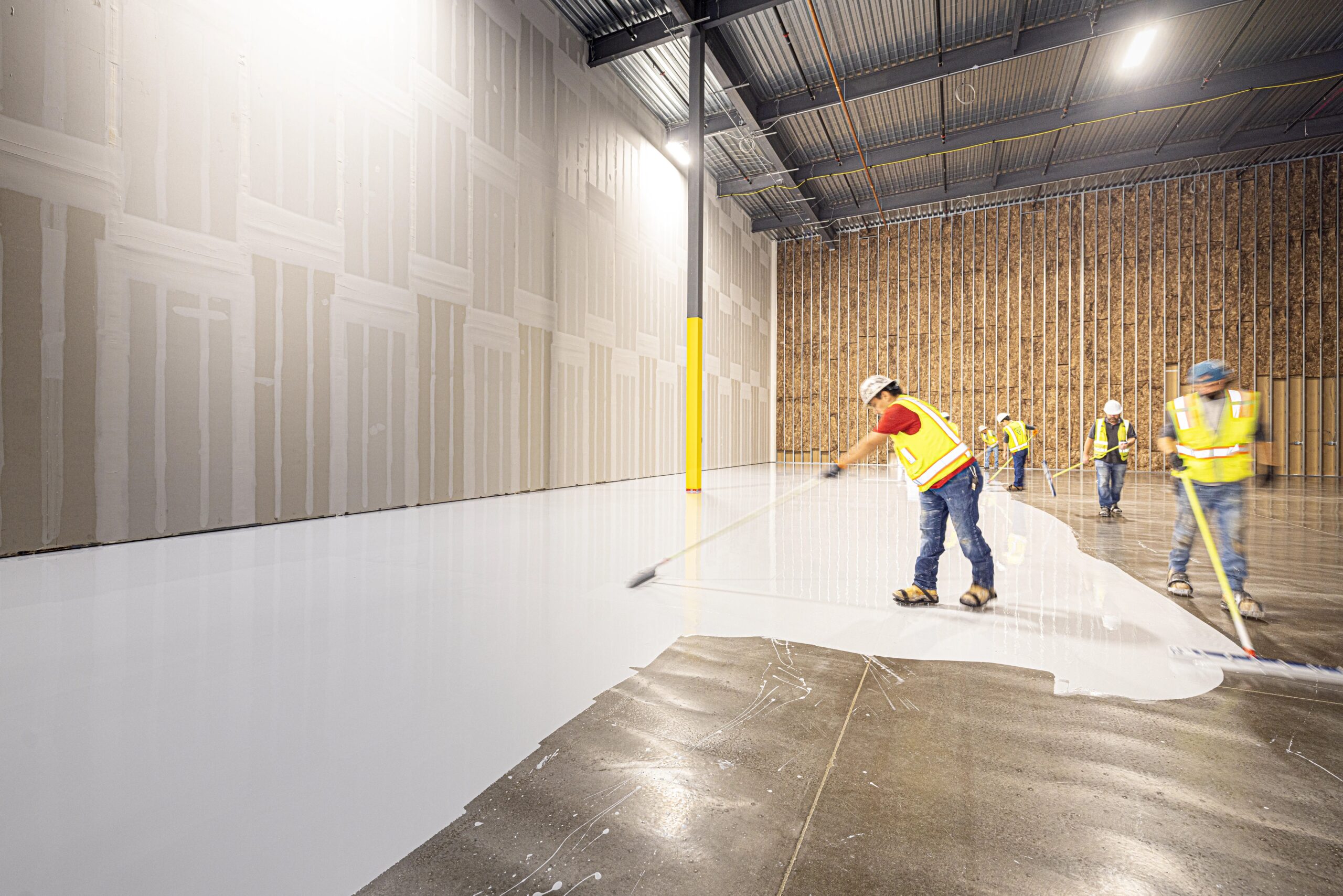 Hantverkare som applicerar ett självutjämnande ytskikt på ett stort betonggolv i en industrilokal under renovering.